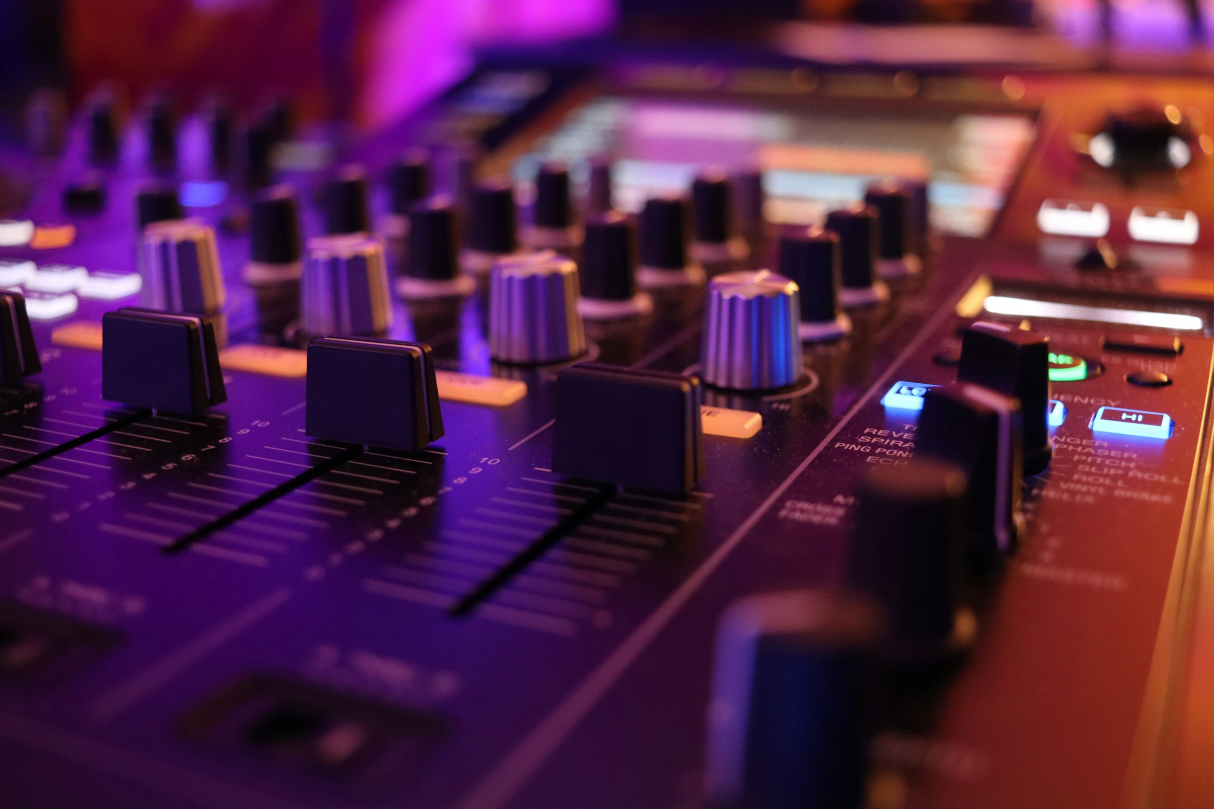 Close-up of a mixing console with knobs, faders, and effect labels under colored lighting.