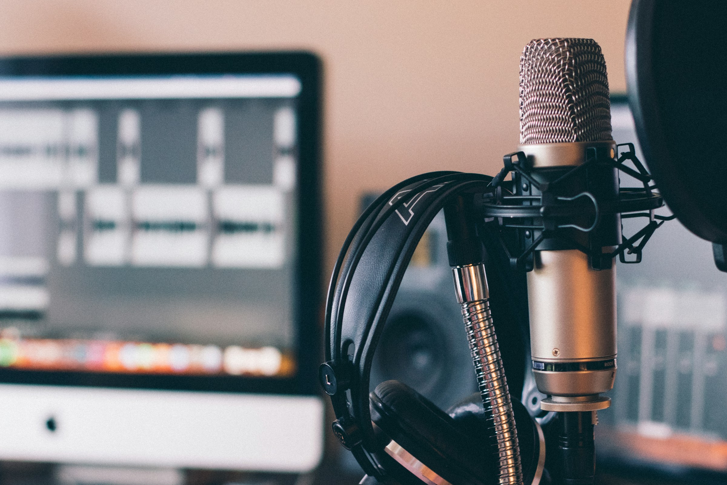 A studio microphone with headphones beside an audio editing workstation showing a waveform.
